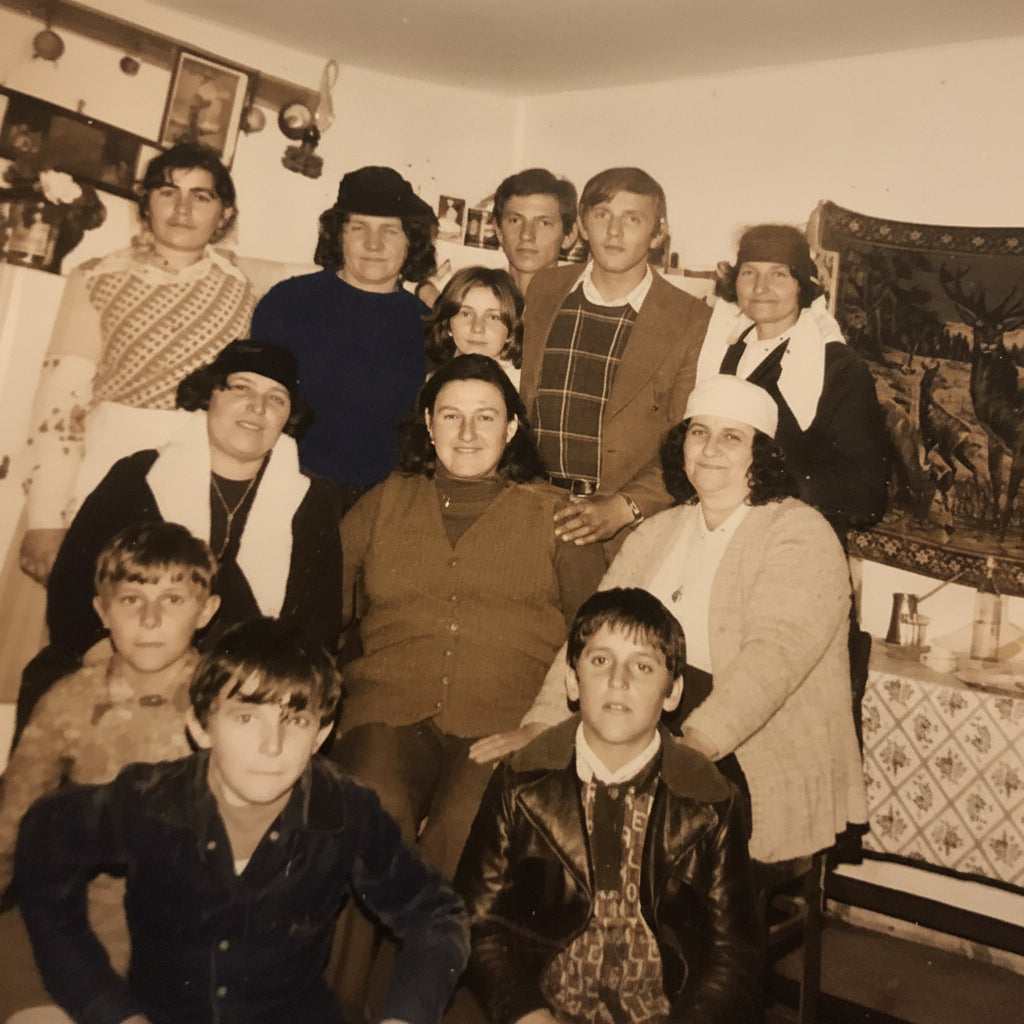 A vintage sepia-toned family portrait showing a group of adults and children posing indoors in what appears to be a home kitchen or dining area, with household decor and tapestries visible in the background.