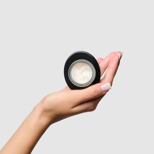 Hand holding a small black container with a white cream inside against a light gray background